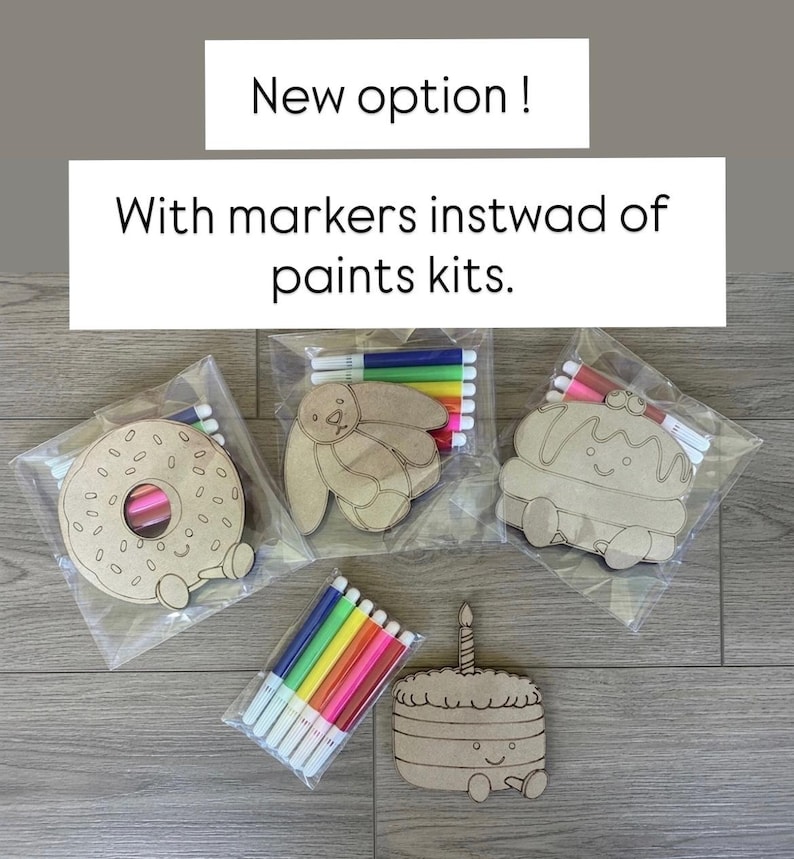 May include: Three clear plastic bags containing light brown wooden cutouts in the shapes of a donut, a rabbit, and a cupcake, each paired with a set of colorful markers. A separate pack of markers and a wooden cake cutout are also visible. Text reads: New option! With markers instwad of paints kits.