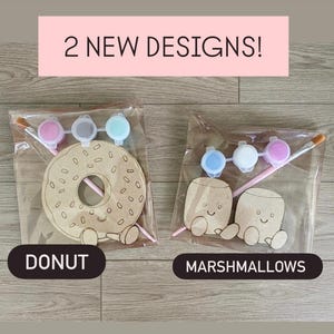 May include: Two craft kits in clear plastic bags. One kit contains a wooden donut cutout with a smiling face, a paintbrush, and three small paint pots. The other kit contains two wooden marshmallow cutouts with smiling faces, a paintbrush, and three paint pots. Text reads 2 NEW DESIGNS!, DONUT, and MARSHMALLOWS.