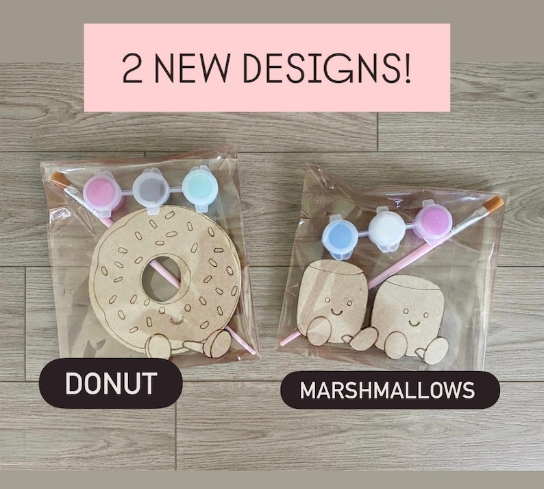 May include: Two craft kits in clear plastic bags. One kit contains a wooden donut cutout with a smiling face, a paintbrush, and three small paint pots. The other kit contains two wooden marshmallow cutouts with smiling faces, a paintbrush, and three paint pots. Text reads 2 NEW DESIGNS!, DONUT, and MARSHMALLOWS.
