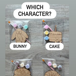 May include: Four individual craft kits in clear plastic bags, each containing a wooden cutout of a bunny, cake, pancake, or croissant, along with a small paintbrush and three small paint pots. The text "WHICH CHARACTER?" appears at the top.