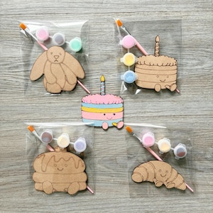 May include: Four clear plastic bags containing wooden cutouts of a rabbit, a cake, a pancake stack, and a croissant, each with a small paintbrush and three paint pots. A single painted cake cutout with a candle sits in the center.