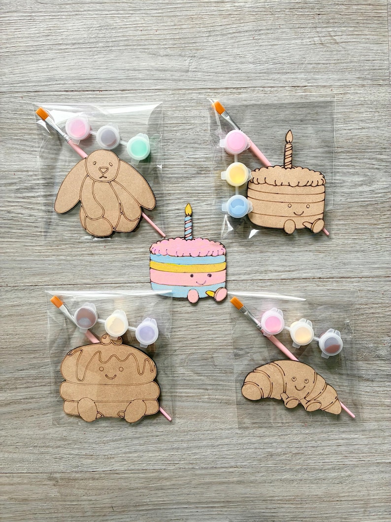 May include: Four clear plastic bags containing wooden cutouts of a rabbit, a cake, a pancake stack, and a croissant, each with a small paintbrush and three paint pots. A single painted cake cutout with a candle sits in the center.