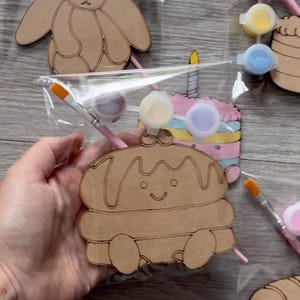 May include: A craft kit featuring a laser-cut wooden shape of a smiling pancake character with syrup, packaged in a clear plastic bag with a small paintbrush and three small circular containers of paint in yellow, purple, and white colors.