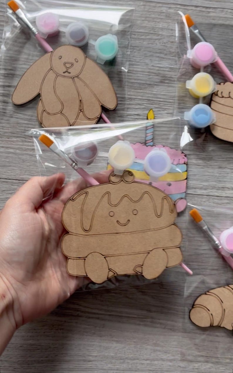 May include: A craft kit featuring a laser-cut wooden shape of a smiling pancake character with syrup, packaged in a clear plastic bag with a small paintbrush and three small circular containers of paint in yellow, purple, and white colors.