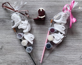 Puede incluir: Dos bolsas de celofán transparente atadas con cintas, cada una con figuras de vaqueros para pintar, botes de pintura y pinceles. También se ve una figura separada de bota de vaquero marrón y blanca. Las figuras incluyen botas de vaquero, caballos y sombreros de vaquero.