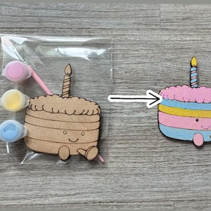 May include: A DIY craft kit featuring a wooden birthday cake cutout with a candle, a paintbrush, and three small paint pots in pink, yellow, and blue. Beside it is a finished, painted version of the same cake in pink, blue, and yellow.