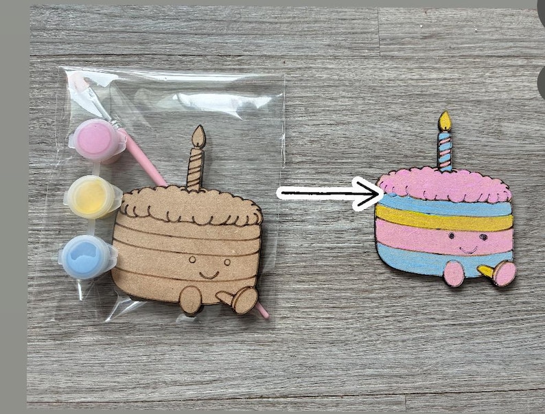 May include: A DIY craft kit featuring a wooden birthday cake cutout with a candle, a paintbrush, and three small paint pots in pink, yellow, and blue. Beside it is a finished, painted version of the same cake in pink, blue, and yellow.
