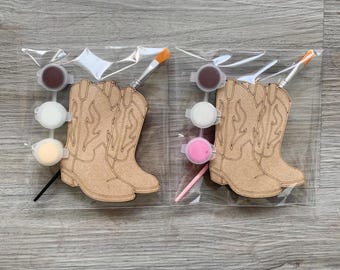 May include: Two paint-your-own cowboy boot kits. Each kit includes a pair of wooden boots, paint pots in brown, white, and yellow, a paintbrush, and a pink stick. The boots are light brown with decorative stitching.