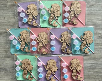 May include: Ten individual craft kits, each sealed in clear plastic, featuring a laser-cut wooden mermaid cutout, a small paintbrush, and three small paint pots containing pink, white, and blue paint, all mounted on a colored cardstock backing.