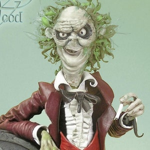 May include: A green-haired, pale-skinned figure with a red bow tie and red pants stands next to a gray tombstone. The figure is holding a small, white object in its right hand. The tombstone has a skull and crossbones design on it. The figure is smiling and has a mischievous look on its face. The text "THE WITCHES OF" is visible in the top left corner of the image.