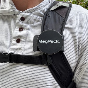 Puede incluir: Una correa de cámara negra con un accesorio magnético redondo negro MagPack. La correa se lleva sobre el hombro de una persona.