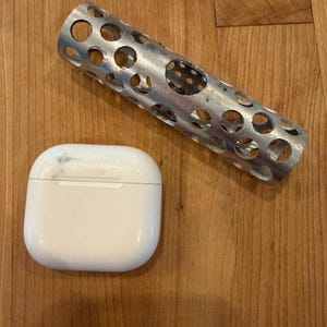 May include: A white, rounded, wireless earbud charging case next to a silver metal tube with multiple circular holes. The items are on a wooden surface.