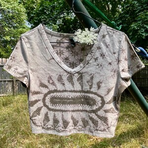May include: A short-sleeve V-neck crop top in a light taupe color featuring a dark brown tie-dye pattern. The design includes a central oval shape surrounded by radiating petal-like motifs, creating a sunburst effect across the front of the garment.