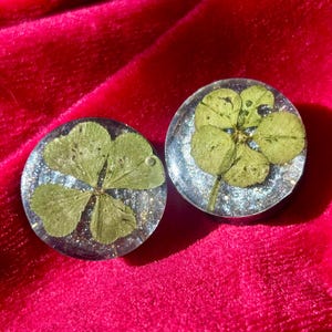May include: Two round, clear resin earrings, each containing a four-leaf clover. The clovers are green, and the resin has a glittery, silver and blue background. The earrings are displayed on a red velvet surface.