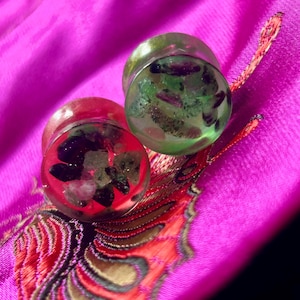 May include: Two clear, round ear plugs with colorful inclusions. One plug has a red base with dark stones, the other has a green base with green and purple stones. The plugs are set against a bright pink fabric.