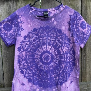 May include: A short-sleeve crew neck t-shirt in deep purple with a bleached-out, light lavender circular doily pattern centered on the front. The garment features a Hanes brand label at the inner neckline and a standard straight hemline.