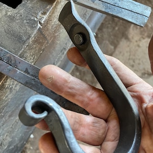 May include: A hand-forged dark gray metal hook with a curved J-shape and a decorative scrolled end. The hook features a flat mounting plate at the top secured by a single hexagonal bolt, showcasing a textured, raw iron finish throughout the piece.