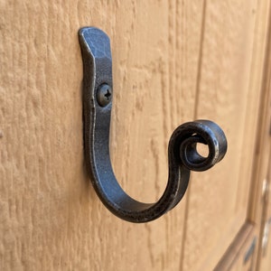 May include: A black metal hook with a decorative scroll design. The hook is mounted on a light brown wall.