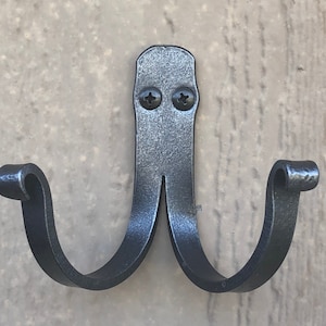 May include: Black metal double hook with a decorative scroll design. The hook is mounted on a wall.