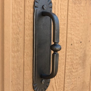May include: A black metal door handle with a decorative design. The handle is attached to a wooden door.