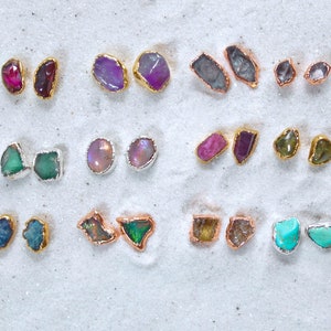 May include: Assortment of gemstone earrings in various colors and shapes, including amethyst, opal, turquoise, and emerald. The earrings are displayed on a white sandy surface.