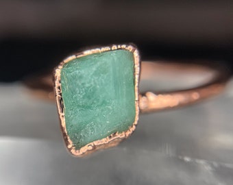 May include: A ring with a raw emerald gemstone set in a rose gold band.