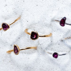 May include: A collection of eight rings with different sized, raw, purple gemstones set in gold and silver bands. The rings are arranged on a white sandy surface.