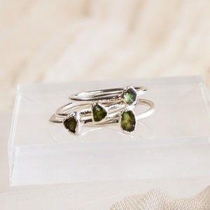 May include: Four silver rings with green gemstone accents stacked on a white display stand.