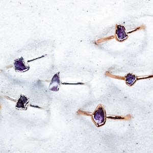 May include: A collection of nine delicate gold rings, each featuring a unique, raw amethyst gemstone. The rings are arranged on a white sandy surface.