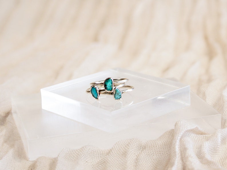 May include: Three silver rings with turquoise stones set in the center of each ring. The rings are stacked on top of each other on a clear acrylic platform.