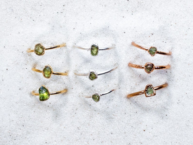 May include: Six gold, silver, and rose gold rings with green gemstone settings. The rings are stacked on a white sandy surface.