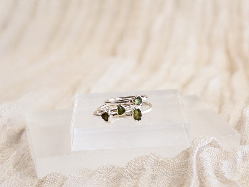 May include: Four silver rings with green gemstone accents stacked on a white display stand.