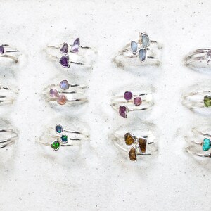 May include: A collection of 12 silver rings with different colored gemstones. Each ring features three gemstones in a cluster. The gemstones are various shades of green, blue, purple, pink, and brown.