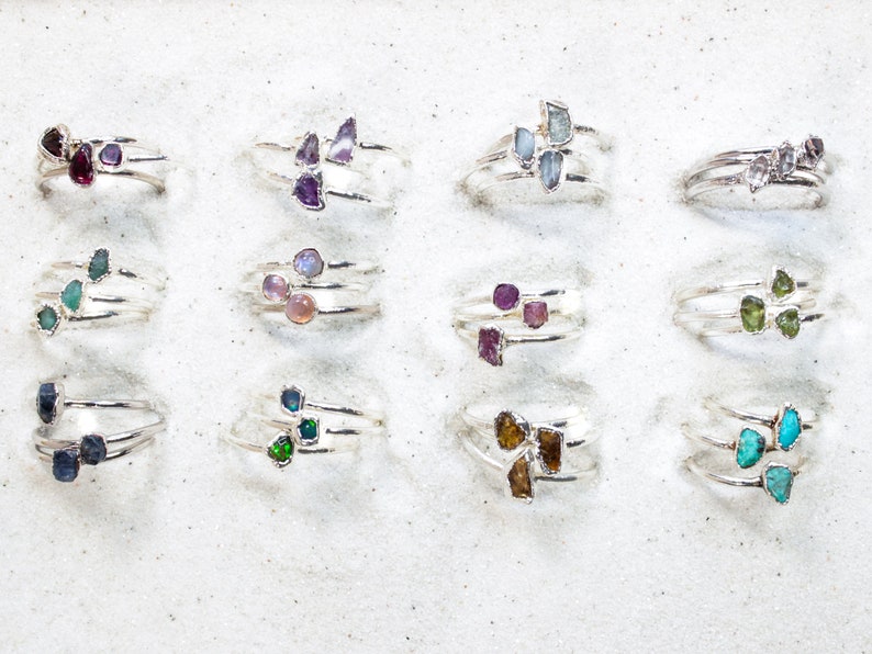 May include: A collection of 12 silver rings with different colored gemstones. Each ring features three gemstones in a cluster. The gemstones are various shades of green, blue, purple, pink, and brown.