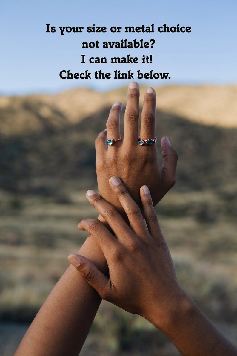 May include: Two hands with rings on the fingers. The rings are made of copper and have green stones. The background is a blurry image of a mountain range.