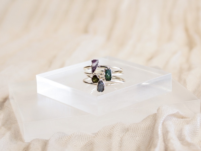 May include: Four silver rings with different colored gemstones. The rings are stacked on top of each other on a white surface.