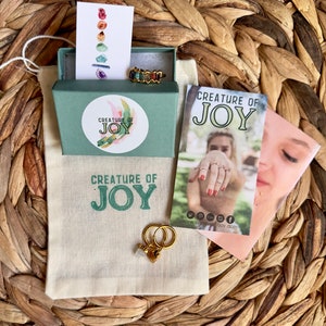 May include: A small green box with a white label that says "Creature of Joy" and a gold ring with colorful stones. The box is sitting on a woven straw surface. There is a white bag with the same logo and a pair of gold hoop earrings with a small crystal charm.