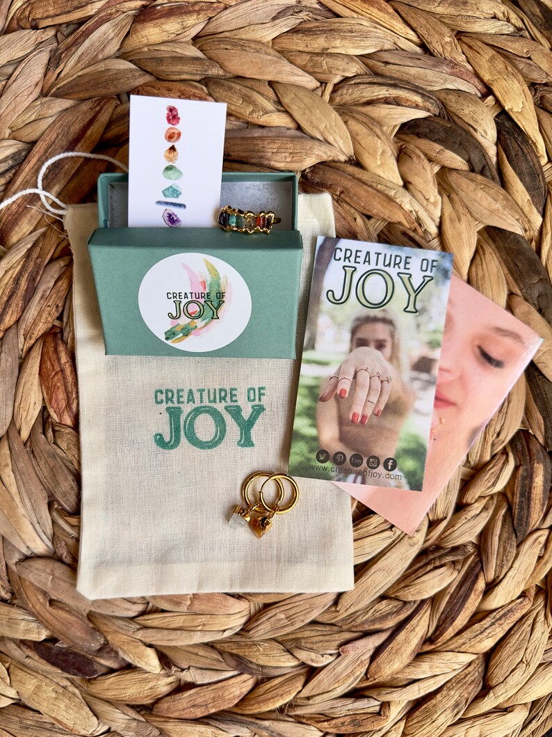 May include: A small green box with a white label that says "Creature of Joy" and a gold ring with colorful stones. The box is sitting on a woven straw surface. There is a white bag with the same logo and a pair of gold hoop earrings with a small crystal charm.