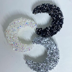 May include: Three crescent-shaped hoop earrings covered in small, reflective crystals. One earring is white with iridescent highlights, one is solid black, and one is a mix of silver and grey tones. Each piece features a dense, textured surface.