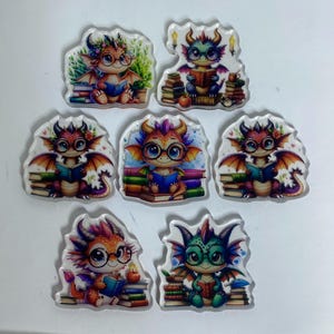 May include: A collection of seven acrylic charms featuring illustrations of small dragons wearing glasses and reading books. Each dragon is depicted in various colors, including orange, green, and purple, surrounded by stacks of books and foliage.