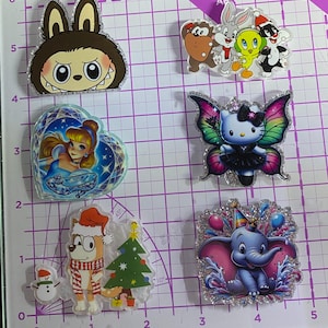 May include: Six acrylic pins featuring various cartoon characters: a brown creature, a group of four animated figures, a princess in a heart frame, a butterfly-winged cat, a dog with a Christmas tree and snowman, and a blue elephant with balloons.