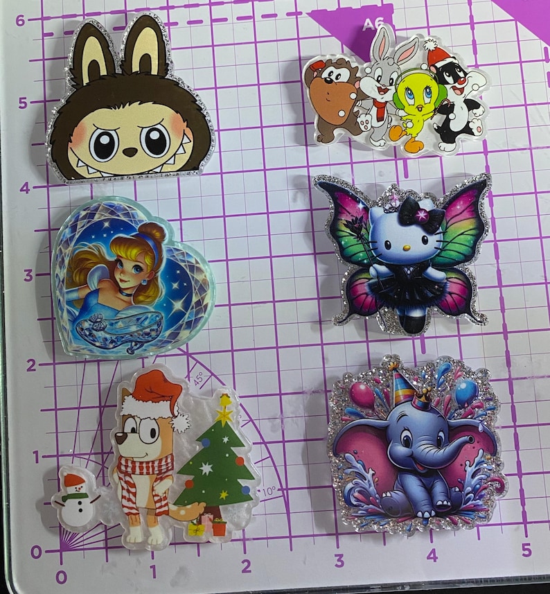 May include: Six acrylic pins featuring various cartoon characters: a brown creature, a group of four animated figures, a princess in a heart frame, a butterfly-winged cat, a dog with a Christmas tree and snowman, and a blue elephant with balloons.