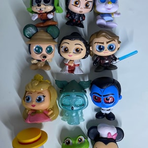 May include: A collection of eleven small stylized vinyl character figurines featuring various animated and cinematic figures. The set includes characters with large eyes, diverse outfits, and accessories like a blue lightsaber, a yellow hat, and a chef hat.