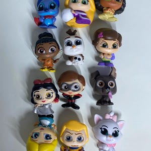 May include: A collection of twelve small, stylized vinyl character figurines featuring various animated figures, including a blue alien, a blonde character with long hair, a dark-haired character with glasses, a snowman, a dog, and a white kitten.