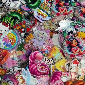 May include: A collection of colourful acrylic charms featuring various cartoon characters, including the Cheshire Cat, Tinker Bell, Hello Kitty, Care Bears, and Moana. Each charm has a glittery border and depicts vibrant, detailed illustrations.