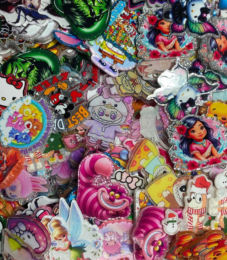 May include: A collection of colourful acrylic charms featuring various cartoon characters, including the Cheshire Cat, Tinker Bell, Hello Kitty, Care Bears, and Moana. Each charm has a glittery border and depicts vibrant, detailed illustrations.