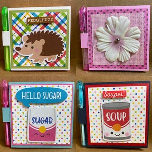 May include: Four small notebooks with attached pens. The notebooks have decorative covers with various designs: a hedgehog with "HEDGEHUGS" text, a white flower, a sugar packet with "HELLO SUGAR!" text, and a soup can with "Souper!" text.