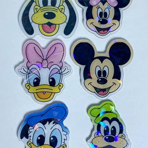 May include: A set of six holographic stickers featuring the faces of Pluto, Minnie Mouse, Daisy Duck, Mickey Mouse, Donald Duck, and Goofy. Each character is depicted with their signature colors, expressions, and features in a die-cut style.