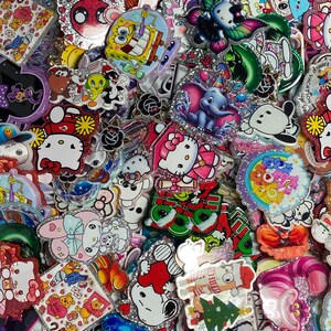 May include: A large collection of colourful acrylic character charms featuring various cartoon figures including Hello Kitty, SpongeBob SquarePants, Dumbo, Snoopy, and Care Bears. Each piece has a glittery finish and distinct printed designs.