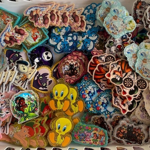 May include: A collection of assorted colourful character stickers featuring various cartoon figures, including Tweety Bird, dalmatians, and fantasy creatures, arranged in a white plastic bin with a grid pattern, showcasing diverse shapes and designs.